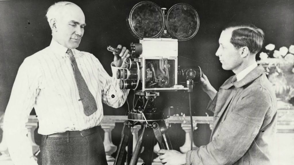 El ingeniero Lee de Forest, pionero de la electrónica, junto a un colaborador mientras trabajan con una cámara de cine antigua equipada con múltiples lentes.