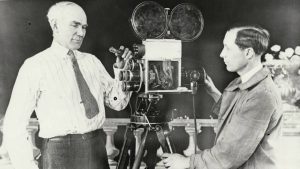 El ingeniero Lee de Forest, pionero de la electrónica, junto a un colaborador mientras trabajan con una cámara de cine antigua equipada con múltiples lentes.