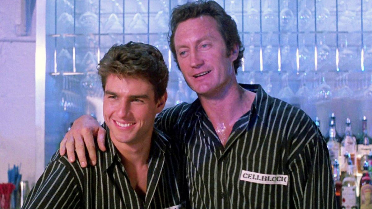 Tom Cruise y Bryan Brown caracterizados como camareros, sonríen juntos detrás de la barra de un bar en una escena de una película de los años ochenta.