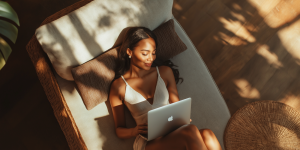 "Mujer joven relajada tumbada en una tumbona, disfrutando de la luz natural mientras trabaja en un ordenador portátil Apple, rodeada de un ambiente acogedor con elementos de madera y mimbre.