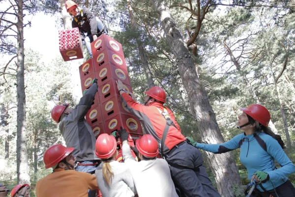 Grupo de personas con cascos de seguridad realizando una actividad de team building en el bosque, apilando cajas mientras una persona en altura coloca otra en la parte superior de la torre.
