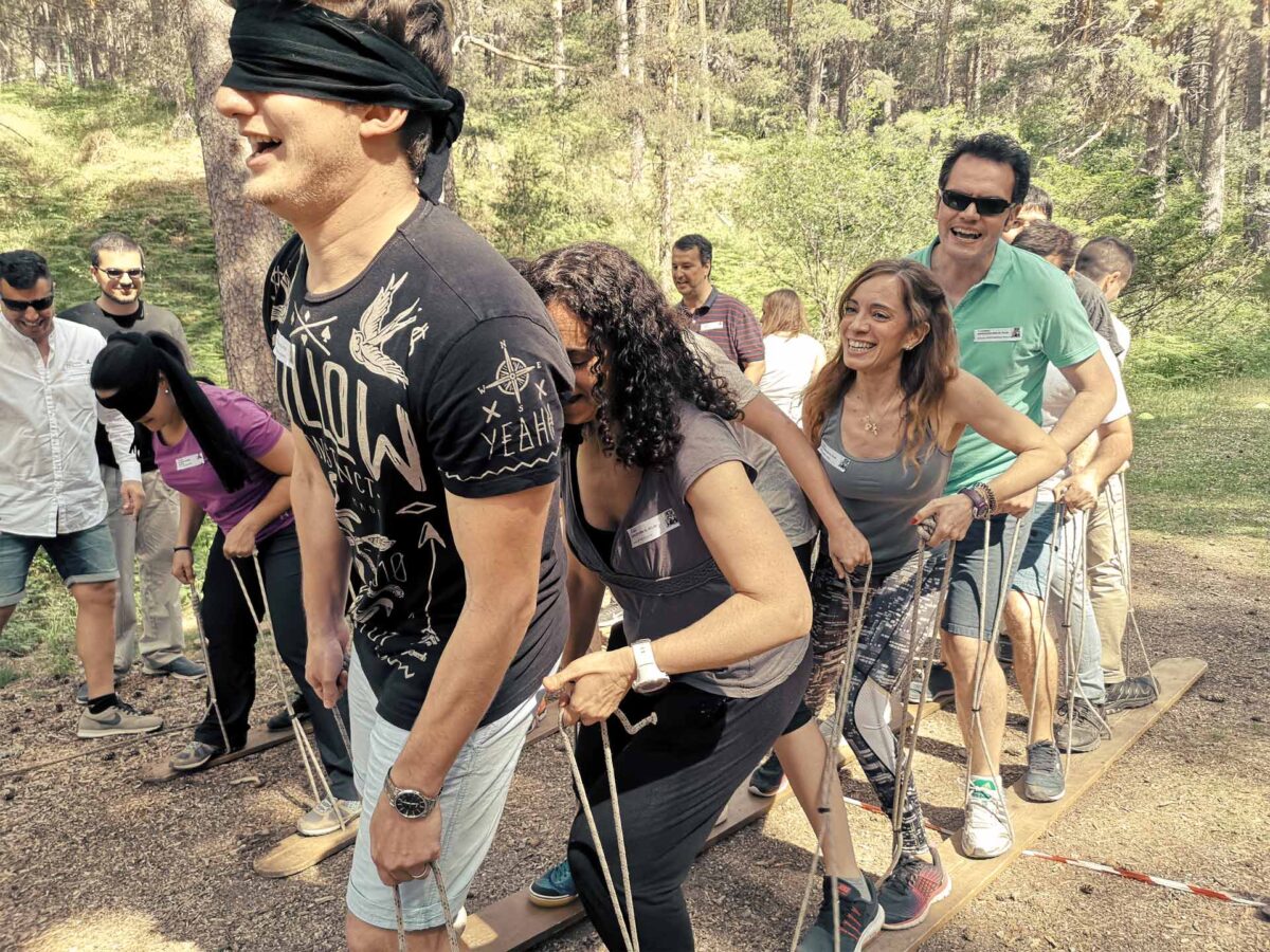 Grupo de adultos participando en una actividad de team building al aire libre en un bosque, caminando coordinados sobre tablas con cuerdas mientras uno de ellos lleva los ojos vendados.