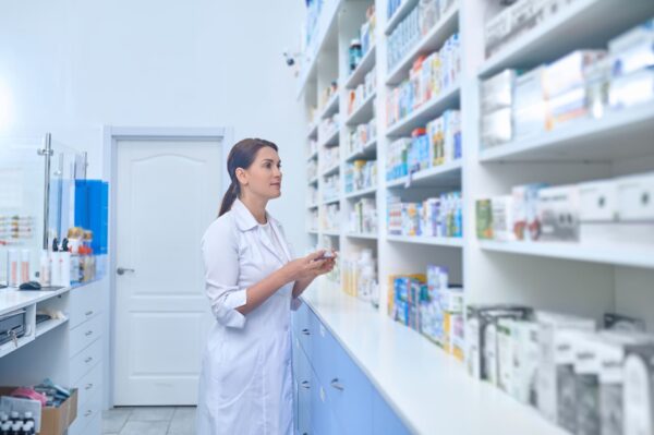 Farmacéutica revisando medicamentos en las estanterías de una farmacia con bata blanca.