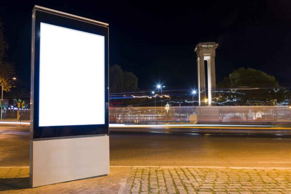 Mupi publicitario iluminado en una calle urbana de noche, con tráfico en movimiento y luces desenfocadas al fondo.
