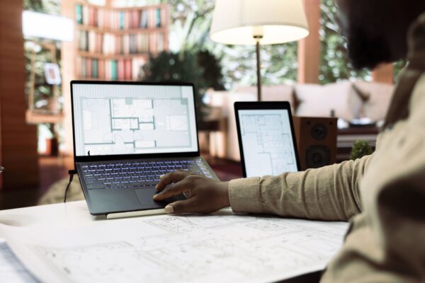 Persona revisando planos arquitectónicos en portátil y tableta sobre una mesa de trabajo en un entorno doméstico o de oficina.