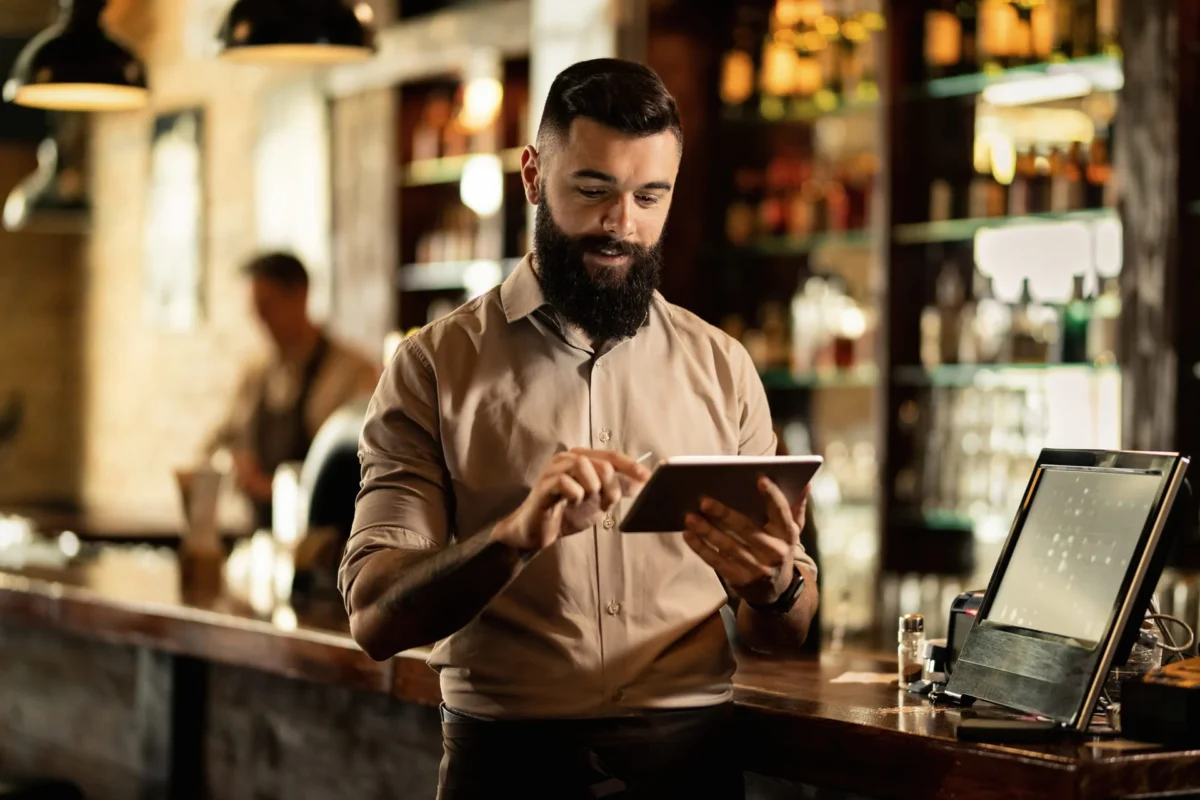 Camarero utilizando una tablet en la barra de un bar, gestionando pedidos en un entorno de hostelería.