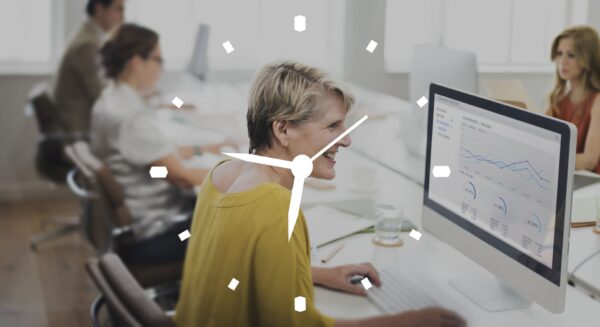 Personas trabajando en una oficina moderna mientras una mujer utiliza un ordenador con gráficos de análisis en pantalla, con un icono de reloj superpuesto que representa gestión del tiempo.