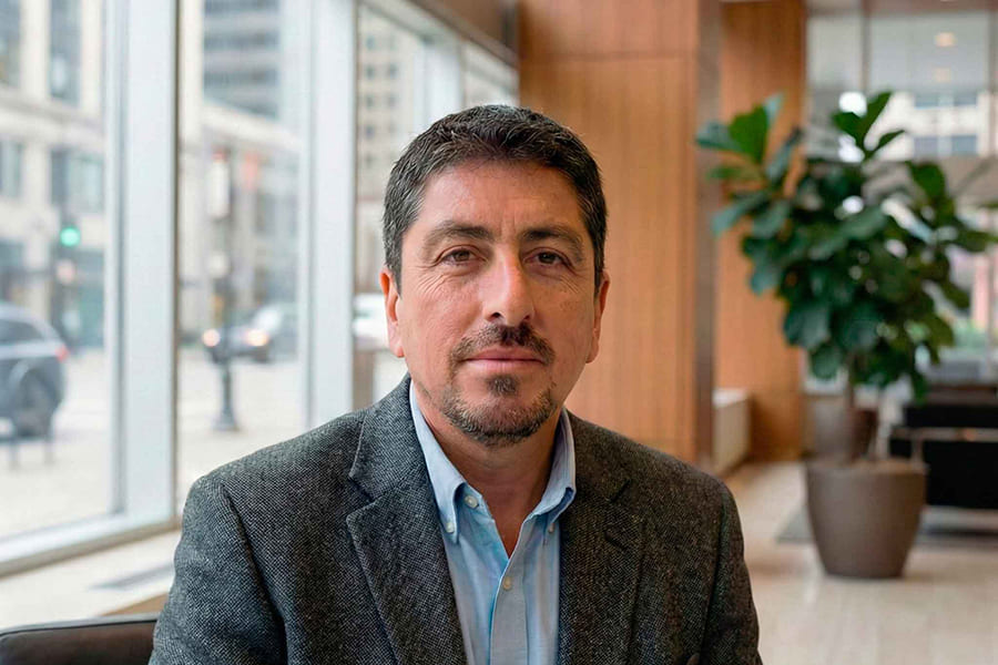 Retrato de hombre con chaqueta gris y camisa azul sentado en una oficina moderna con ventanales y plantas al fondo.