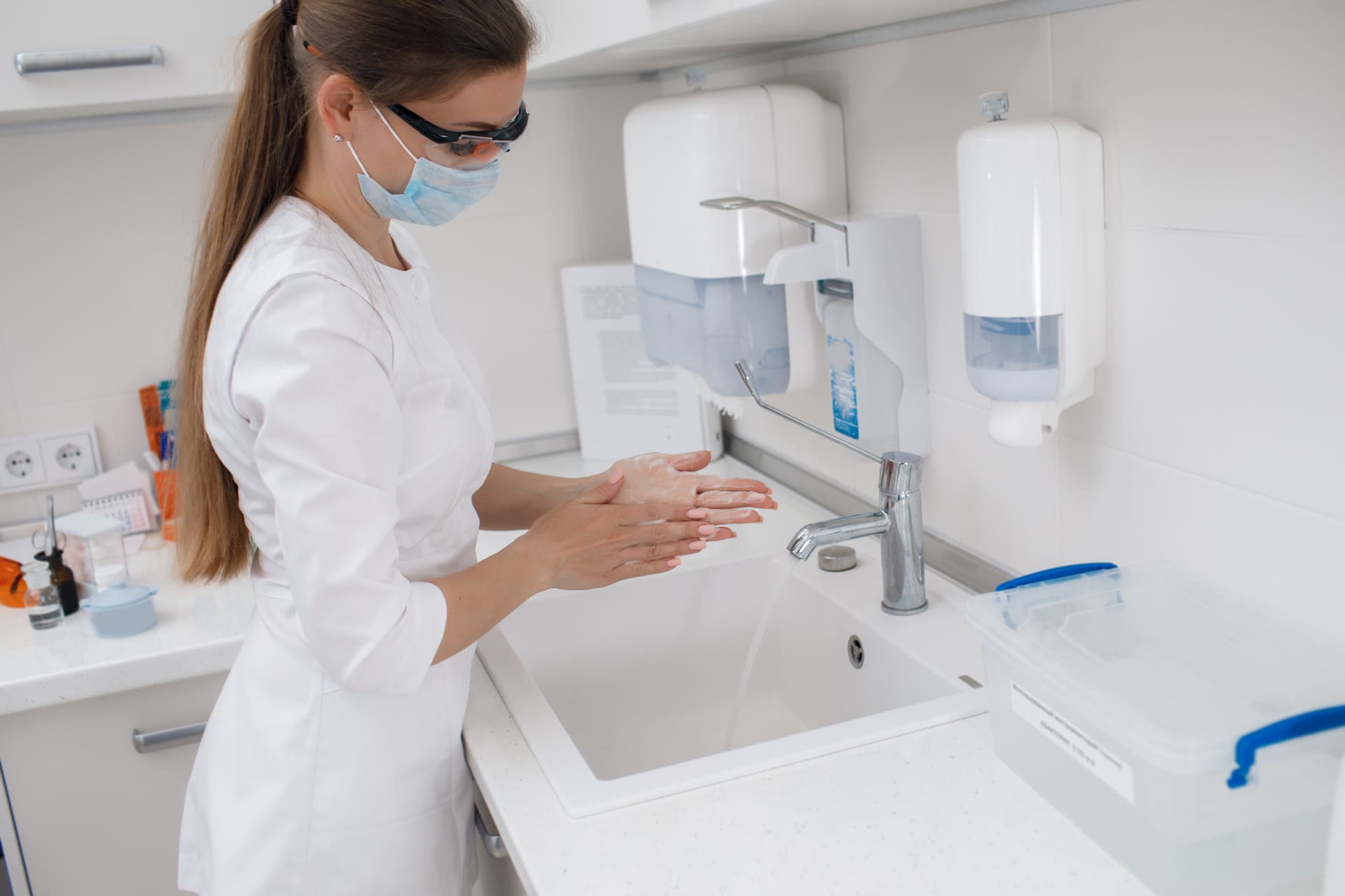 Profesional sanitaria con mascarilla y gafas de protección lavándose las manos en un entorno clínico, siguiendo protocolos de higiene y seguridad sanitaria.