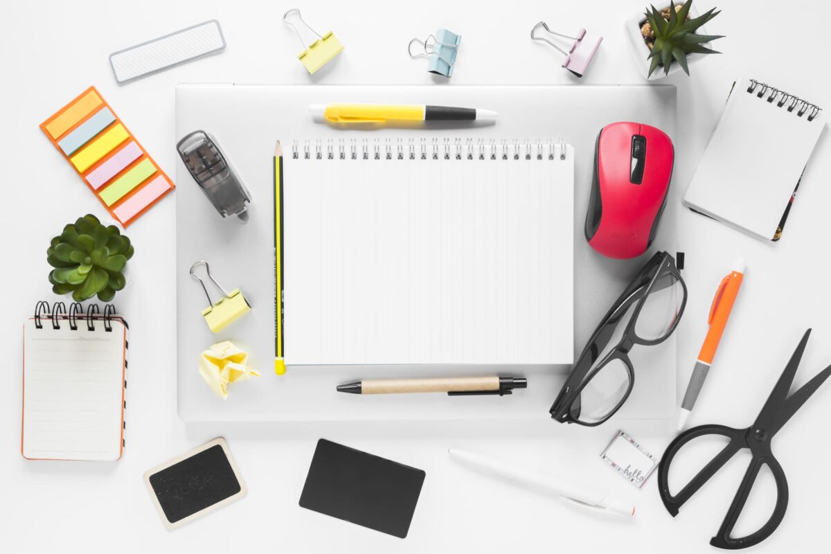 Vista cenital de un escritorio de oficina con cuaderno en blanco, material de papelería, ratón, gafas, notas adhesivas y accesorios de trabajo organizados sobre una superficie blanca.