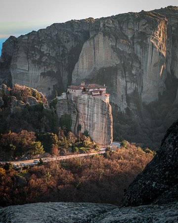 El Monasterio de Rousanou edificado sobre una imponente formación rocosa vertical en Meteora, Grecia, rodeado de vegetación durante el atardecer.