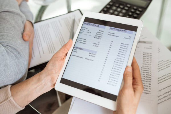 Persona sosteniendo una tablet con una hoja de cálculo de inversiones y gastos en la pantalla, con una calculadora y documentos en papel de fondo.