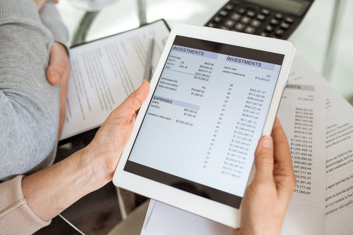 Persona sosteniendo una tablet con una hoja de cálculo de inversiones y gastos en la pantalla, con una calculadora y documentos en papel de fondo.