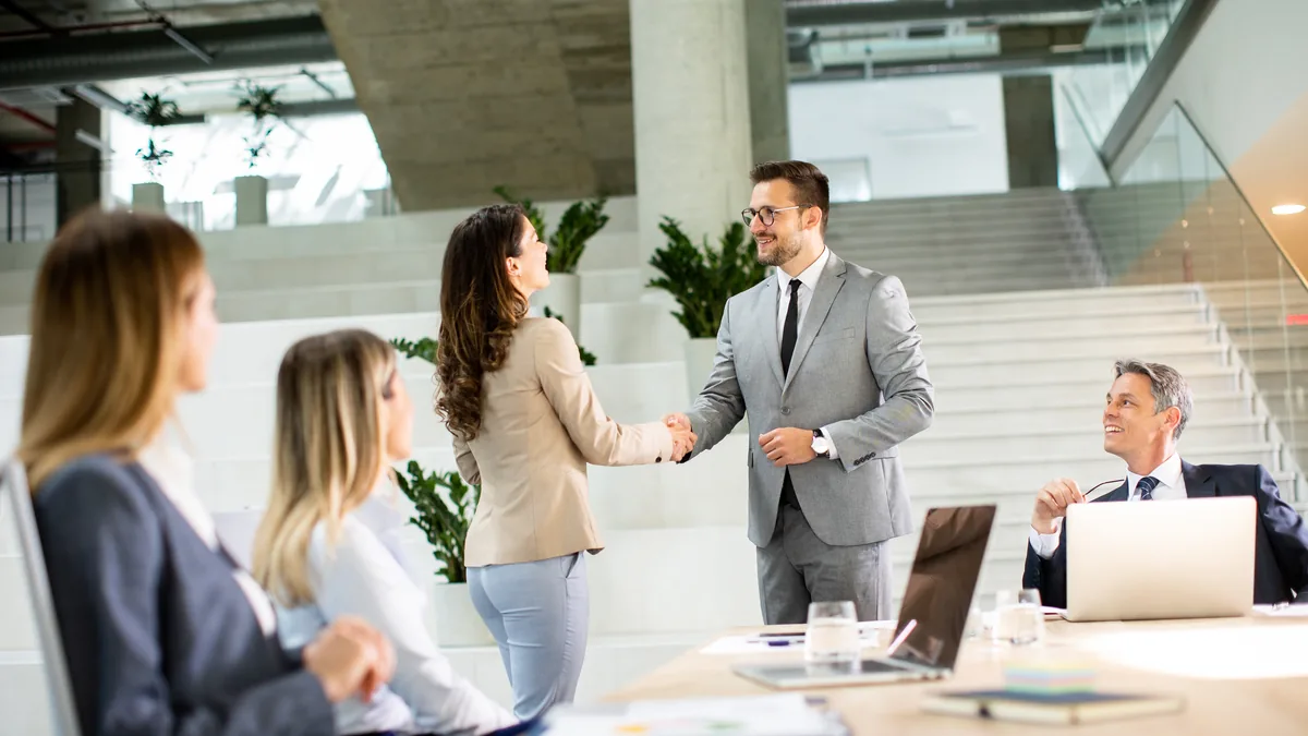 Dos profesionales se dan la mano de pie junto a una mesa de reuniones con portatiles, mientras el resto del equipo observa sentado en una oficina moderna.