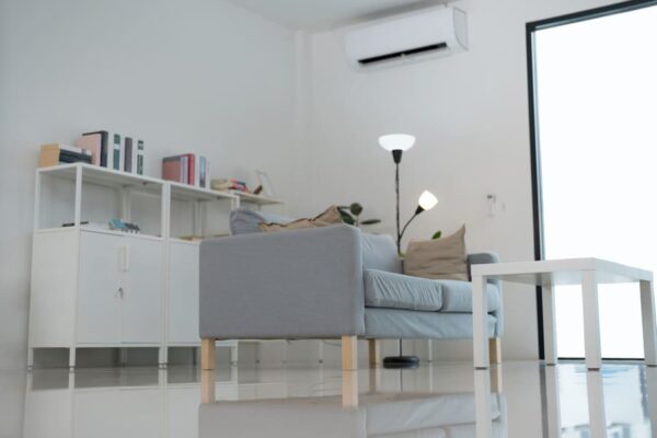 Aire acondicionado split montado en la pared de un salón minimalista, junto a un sofá gris, estantería blanca con libros, lámpara de pie y puerta de cristal a la terraza.