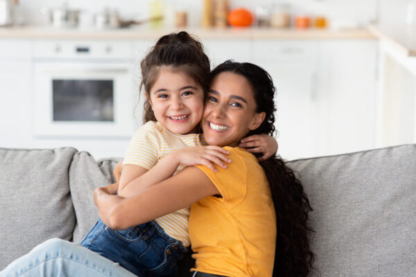 Madre e hija abrazadas y sonrientes en un sofá gris, con una cocina de fondo, en un momento cálido en casa