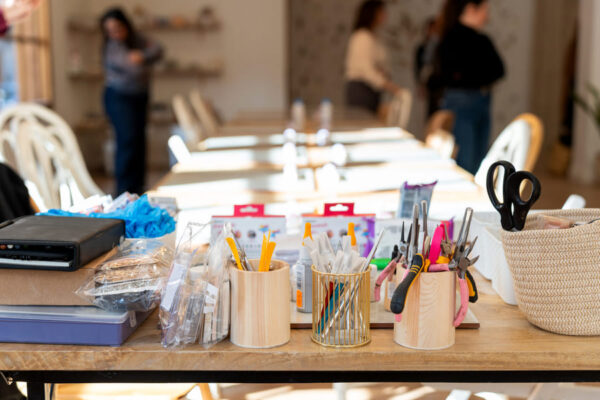 Mesa larga de taller con botes de herramientas de manualidades —tijeras, alicates y bolígrafos—, blísters de material y una cesta de mimbre en primer plano, con varias personas conversando desenfocadas al fondo de la sala