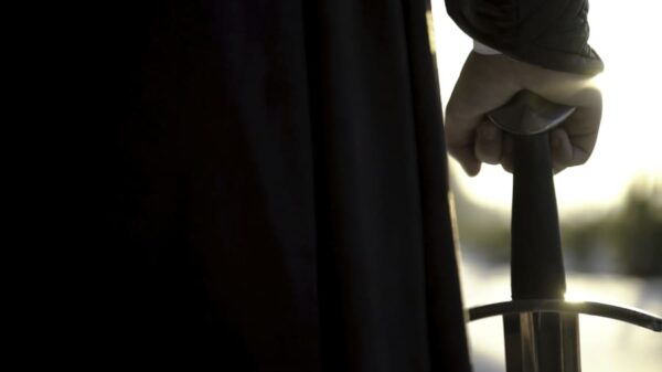 Mano empuñando la cazoleta de una espada de reproducción histórica junto a una cortina oscura, a contraluz