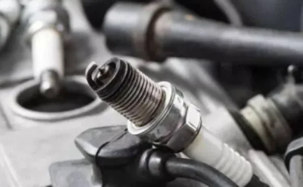 Bujía de encendido en primer plano sobre el motor de un vehículo, junto a cables y componentes mecánicos.