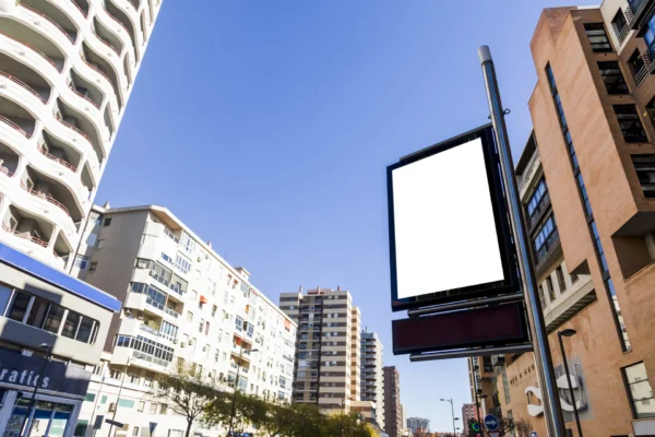 Tótem publicitario con pantalla en blanco instalado en una calle urbana rodeada de edificios bajo un cielo despejado