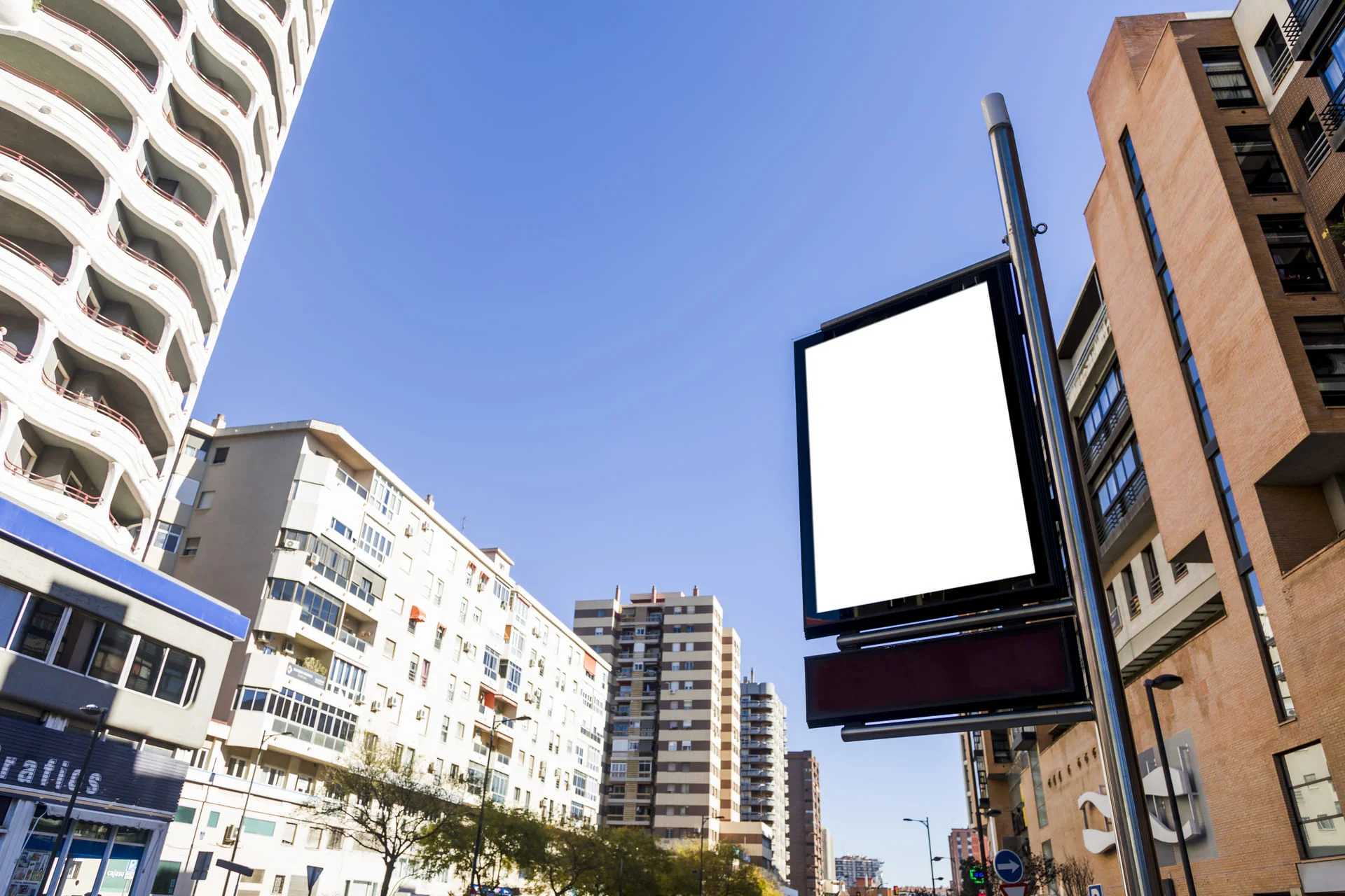 Tótem publicitario con pantalla en blanco instalado en una calle urbana rodeada de edificios bajo un cielo despejado