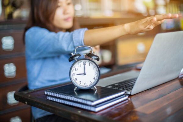 Mujer estirando los brazos frente a un portátil, con un reloj y cuadernos sobre la mesa.
